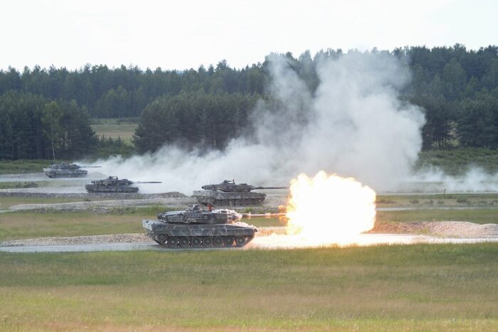 Ein Feld, dahinter Wald, mehrere Panzer hintereinander auf dem Feld, der erste schießt gerade, aus der Mündung kommt Feuer.