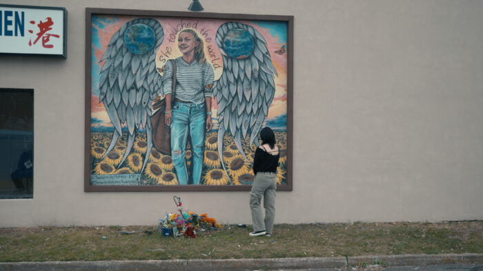 Man sieht ein Mural, das an die ermordete Gabby Petito erinnert, davor liegen Blumen und eine Person steht da und schaut auf das Mural.