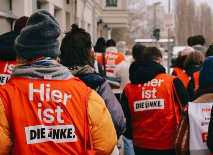 Straßenfoto von einer Gruppe von Menschen, die auf einem Gehweg neben einem Haus laufen. Sie tragen rote Jacken, auf dem steht: "Hier ist die Linke"