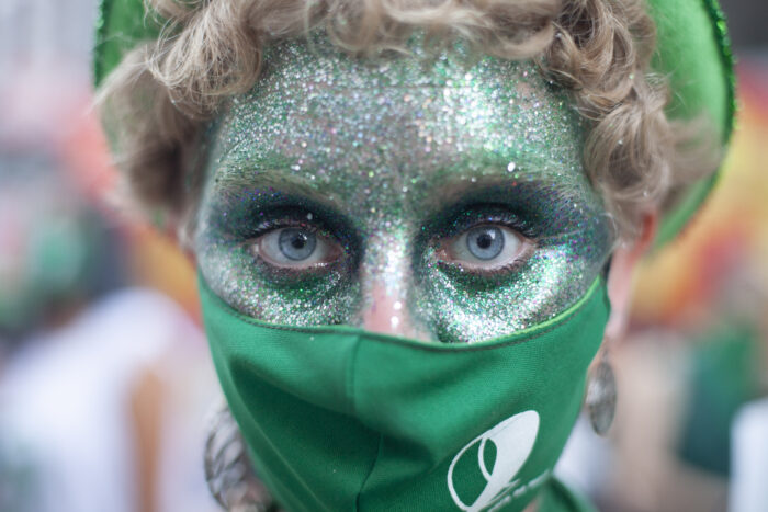 Eine Person mit grünem Glitzer im Gesicht und einer grünen Maske vor dem Mund, die in die Kamera blickt.