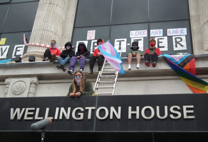 Aktivist*innen mit Trans-Pride-Flaggen auf dem Gebäude des nationalen Sicherheitsdienstes Großbritanniens (NHS).