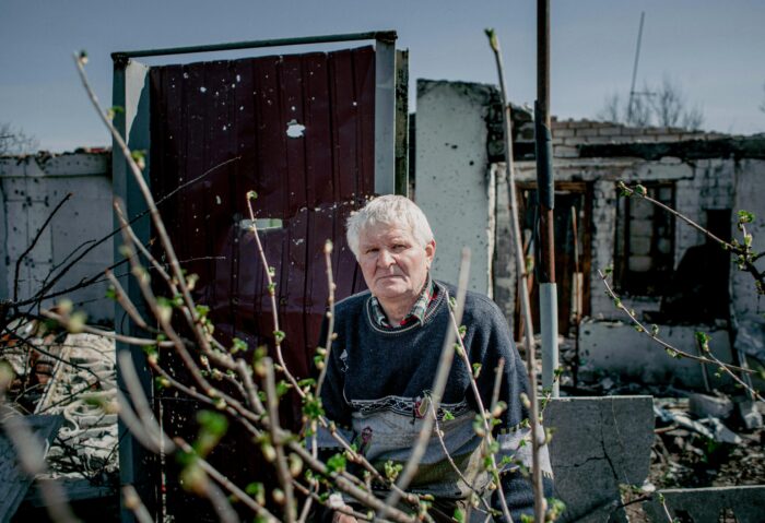 Ein alter Mann vor einem zerstörten, zerschossenen Haus blickt ernst in die Kamera